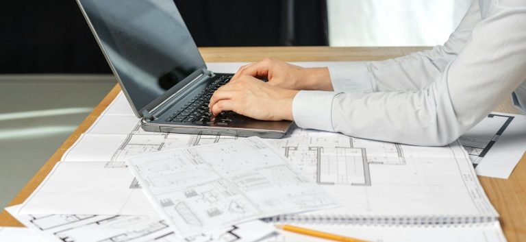 Audiovisual installation Wales Person typing on a laptop, surrounded by architectural plans on a table.