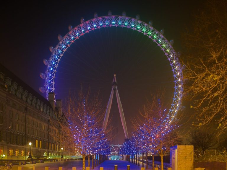 The London Eye illuminated at night, surrounded by colourful lights and trees.