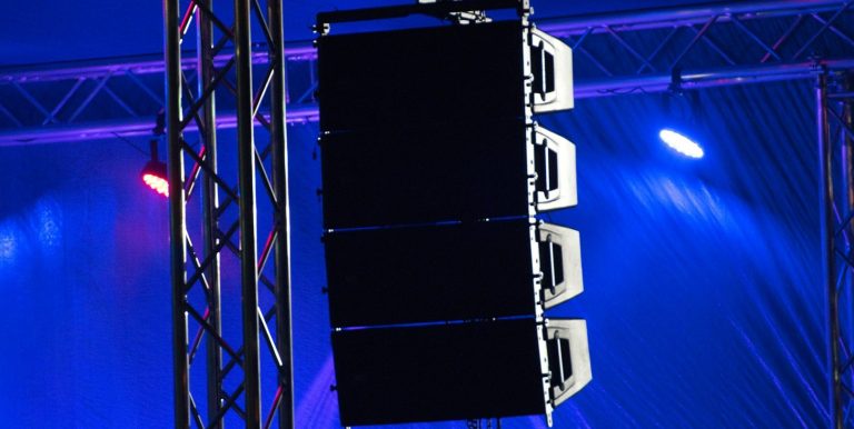 Four stacked speakers suspended in a concert setting with colourful stage lighting.