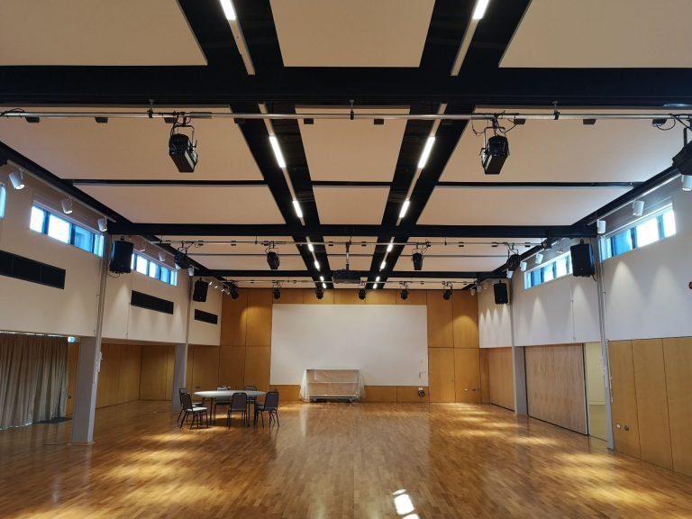 Spacious indoor hall with wooden flooring, large windows, and minimal furnishings.