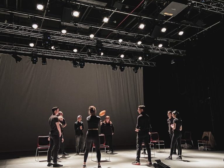 A group of performers on stage rehearsing with chairs and stage lighting.